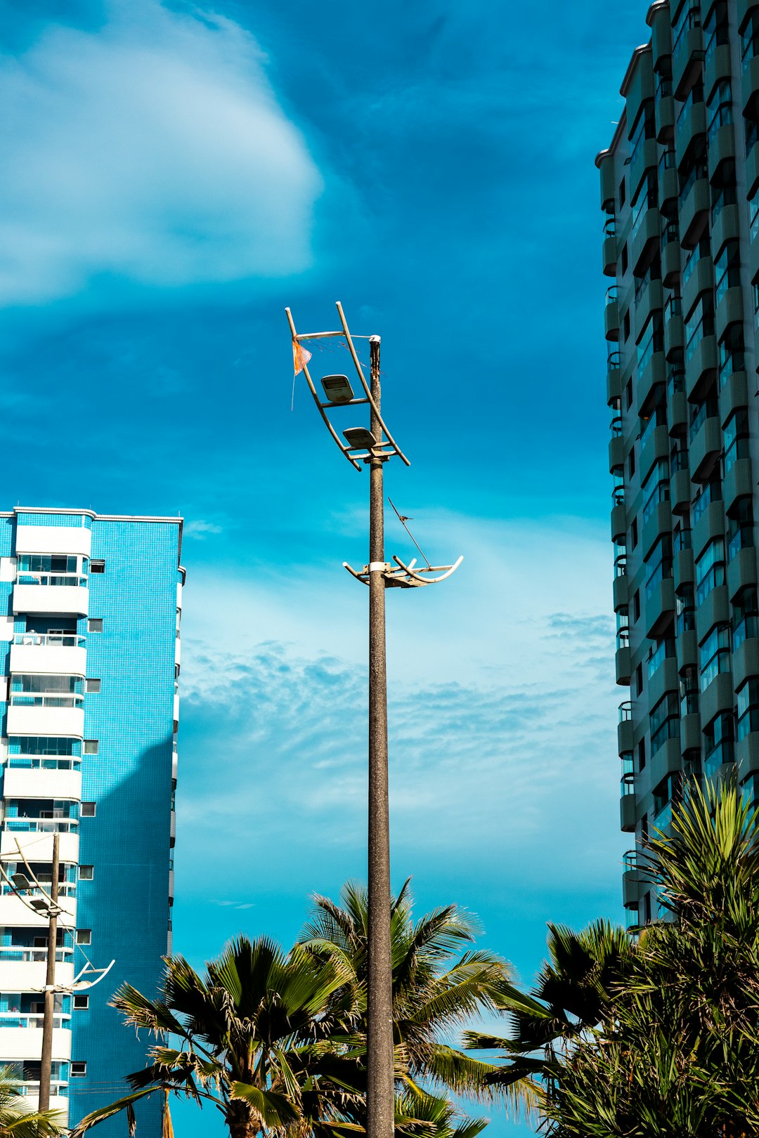 A tall, weathered lamppost stands between two modern apartment buildings under a bright blue sky with scattered clouds. Palm trees frame the bottom of the image, adding a tropical feel to the urban landscape.